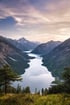 Ein langer, schmaler See schlängelt sich durch ein Tal, umgeben von hohen Bergen und bewaldeten Hängen unter einem bewölkten Himmel.