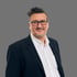 Ralph Fritsch, professionelles Business-Porträt eines Mannes mit Brille und dunklem Anzug vor neutralem grauen Hintergrund.