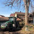 Ein grüner Traktor mit Greifarm arbeitet an einem großen Baumstamm in einem Garten vor Wohnhäusern.