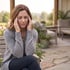 A middle-aged woman sits on an outdoor bench with eyes closed, rubbing her temples as if experiencing a headache or migraine.