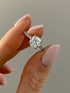 Ein runder Diamant-Verlobungsring aus Weißgold mit funkelndem Besatz auf der Ringschiene wird zwischen zwei Fingern gehalten.