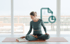 Eine Frau sitzt im Schneidersitz auf einer Yogamatte und liest ein Buch vor einer Fensterfront mit Stadtblick.
