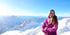 Frau im pinkfarbenen Anorak steht lächelnd vor einem weiten Panorama schneebedeckter Berge unter blauem Himmel.