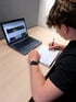 Junger Mann mit Smartwatch schreibt mit Stift in Notizbuch, daneben ein MacBook mit Website.