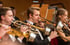 Drei Musiker in Smoking und Abendkleid spielen Posaune und Trompete in einem Orchester.