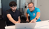 Zwei Männer blicken auf einen Laptop; der jüngere links mit Bart, der ältere rechts in blauem T-Shirt zeigt auf den Bildschirm.