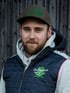 Ein bärtiger Mann mit Kappe und Weste der Firma Da Holzknecht Baumpflege vor einer Holzwand.