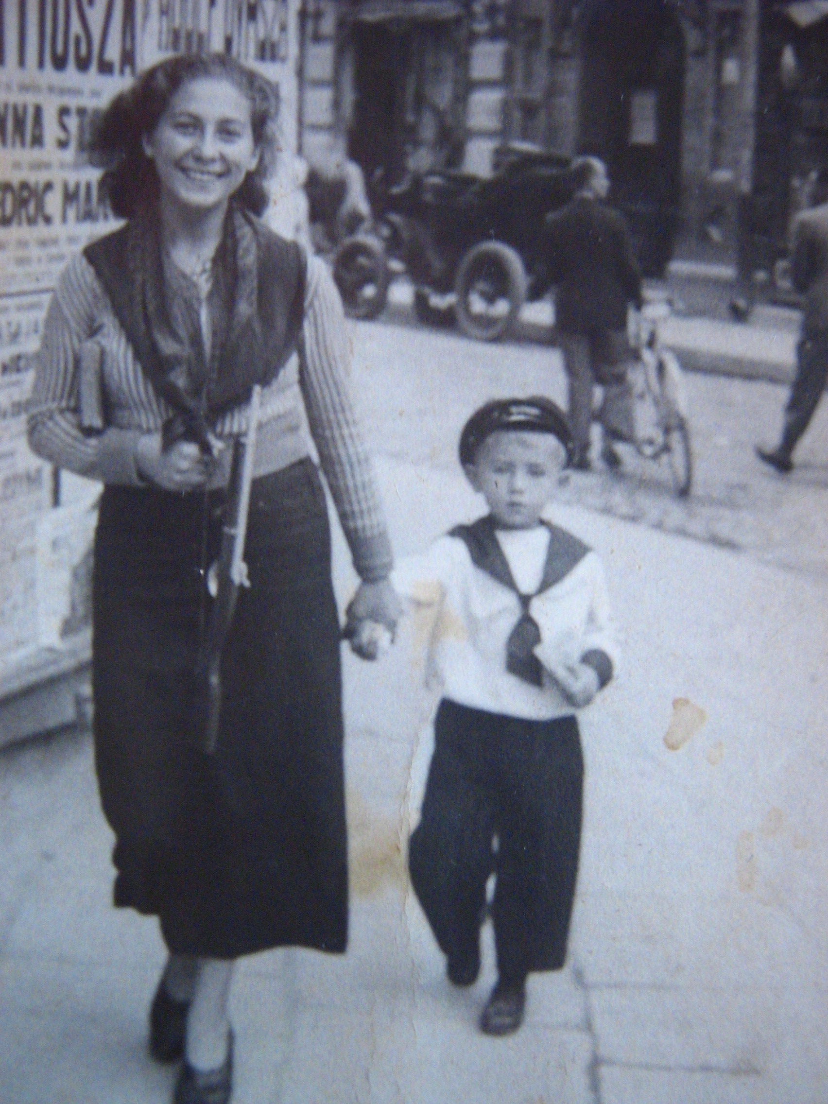Foto en blanco y negro de Bela Świ, hermana del medio de Hirsz, caminando por una calle del centro de Grodno tomada de la mano de un niño pequeño vestido con traje de marinero. Bela mira a cámara sonriendo, lleva una blusa de rayas finas, falda larga oscura, un pañuelo cruzado al cuello y libros bajo el brazo izquierdo. Detrás se ve un panel de afiches con el cartel grande de la película «KATIUSZA» (Anna Sten, Fredric March), automóviles Ford de los años 30 estacionados en doble fila y peatones por la vereda. La calle está perfectamente pavimentada — sin escombros, sin daños visibles.