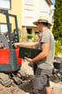 Jens Gandyra mit Strohhut und Mappe zeigt bei einer Gartenbau-Baustelle auf einen roten Minibagger vor einem Wohnhaus.