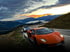 Oranger Lamborghini und silberner Sportwagen auf Bergstraße bei Sonnenuntergang, Berge und Wolken im Hintergrund.