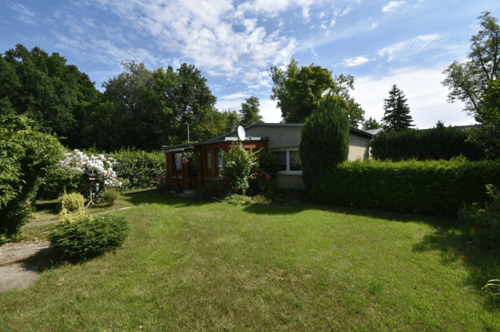 Kleines Haus mit Veranda und Satellitenschüssel, umgeben von grünen Büschen, Bäumen und Rasen unter blauem Himmel.