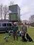 Zwei Männer und zwei Hunde stehen neben einem grünen, hohen mobilen Hochsitz auf einem Anhänger.