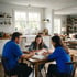 A man and woman sit at a wooden dining table helping a young girl with her schoolwork in a bright, modern kitchen.