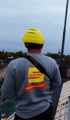 Rückenansicht eines Arbeiters der Schiffer Bedachungen mit gelbem Helm und grauem Sweatshirt mit „SCHIFFER BEDACHUNGEN“ auf dem Dach, der einen großen Kran fernsteuert.