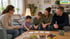 Eine Beraterin sitzt einer Familie mit zwei Kindern am Wohnzimmertisch gegenüber, auf dem Karten und Spielzeug liegen.