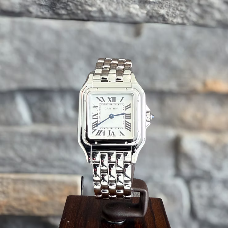 Silberne Cartier Panthère Uhr mit römischen Ziffern und blauem Cabochon, auf Holzständer vor Steinwand.