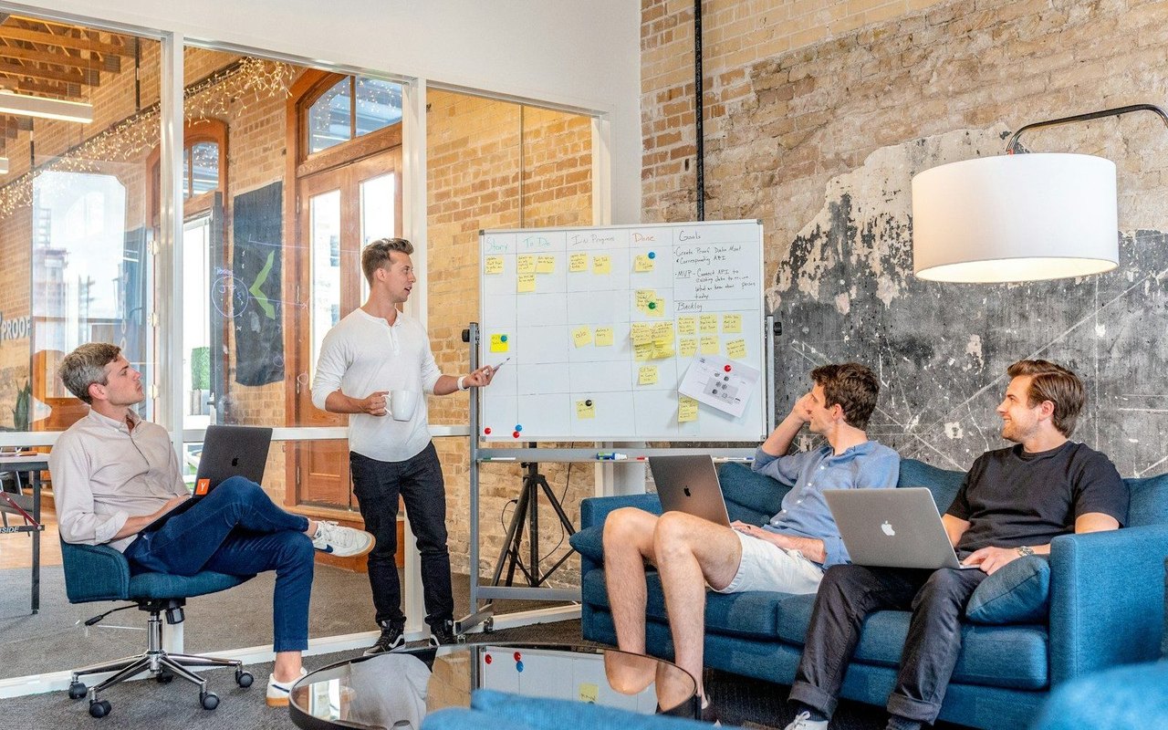 Vier Kollegen mit MacBooks in einer Besprechung vor einem Whiteboard mit Haftnotizen