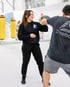 Lächelnde Trainerin im schwarzen Hoodie hält Pratzen für einen Boxer im Boxclub BACKYARD mit gelben Boxsäcken.