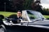 Ein Mann im Smoking sitzt in einem schwarzen Oldtimer-Cabriolet vor einer grünen Wiese und Bäumen.