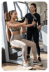 Zwei Frauen im Fitnessstudio: Eine trainiert an einer Brustpresse, die andere steht daneben und gibt Anweisungen.