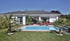 Moderner weißer Bungalow mit Swimmingpool, Terrasse, roten Liegestühlen und Palmen im Garten unter blauem Himmel.