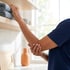 A person in a blue shirt reaches for a folded towel on a wooden shelf while holding their elbow in discomfort.