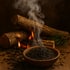 A wooden bowl of dark tea leaves with smoke rising, in front of burning logs.