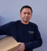 Mann in blauem Pullover mit "Transporte & Umzüge FLEISCHER" Text, hält einen Karton.