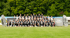 Große Gruppe Kinder und Erwachsene in Sportkleidung auf einem grünen Feld vor Bäumen, mit zwei aufblasbaren Figuren und zwei Schildern.
