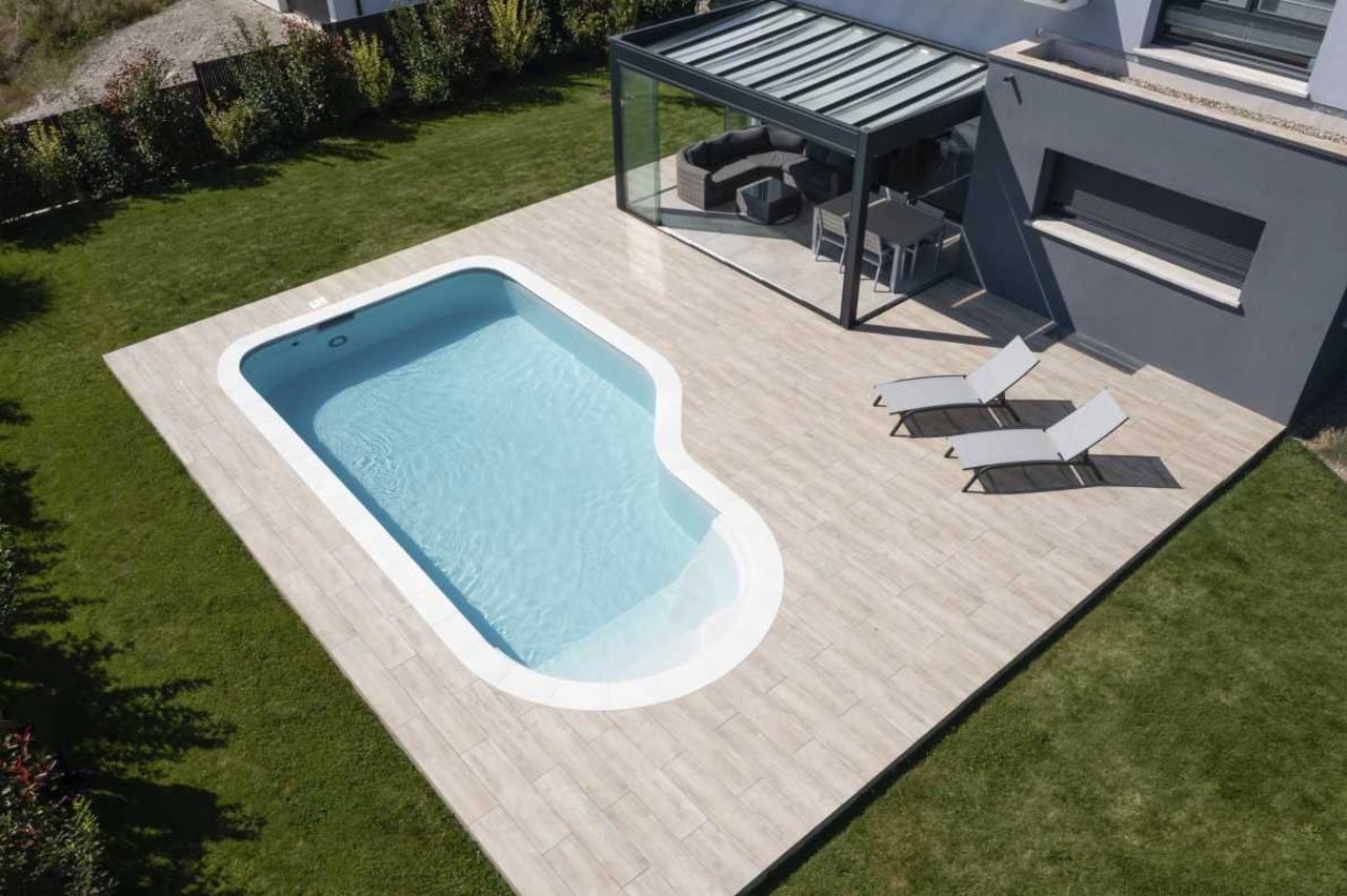 Nierenförmiger Pool mit hellem Wasser auf einer Terrasse, daneben zwei Sonnenliegen und ein Gartenhaus.