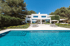 Moderne weiße Villa mit großem Pool und Terrasse, umgeben von grünen Bäumen unter blauem Himmel.