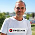 Ein lächelnder Mann im weißen T-Shirt mit dem Logo-Schriftzug UNGLEHRT vor einem sonnigen, unscharfen Hintergrund.