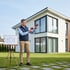 Mann mit Wärmebildkamera vor einem modernen Einfamilienhaus neben einem Stativ mit Bauplan auf einer Rasenfläche.