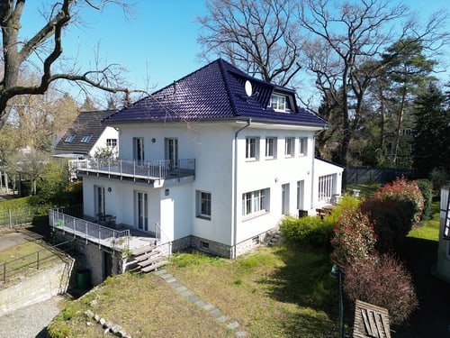 Weißes dreistöckiges Haus mit blauem Dach und Solarpaneelen, umgeben von Bäumen und einem Garten.