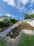 Moderne Außentreppe aus Betonblöcken führt durch einen bepflanzten Hang mit Natursteinmauer und Rindenmulch zum Haus.