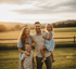 Eine lächelnde Familie mit zwei Kindern steht in einem sonnigen Feld mit einem Holzzaun und Bergen im Hintergrund.