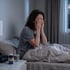 A woman sits in bed at 3:47 AM rubbing her eyes next to a bottle of melatonin and a glass of water.