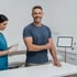 smiling man in a blue t-shirt stands next to a white counter while a nurse in blue scrubs prepares a syringe for low testosterone treatment