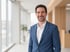 Mann in blauem Anzug und weißem Hemd lächelt vor einem hellen Büroflur.
