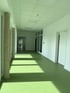 Heller Flur mit grünem Boden, weißen Wänden, Fenstern und Säulen, durch die Sonnenlicht Streifen auf den Boden wirft.