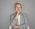 Eine Frau mit kurzen blonden Haaren, einem grauen Blazer und einem weißen Hemd lächelt und stützt das Kinn auf die Hand.