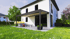 Moderne weiße Hausfassade mit Pergola, grauen Gartenmöbeln auf Terrasse und grüner Rasenfläche.