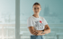 Lächelnde Frau mit Brille und T-Shirt mit der Aufschrift „MY HEART BEATS TAX“ vor verschwommener Stadtkulisse mit dem Kölner Dom.