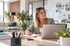 Lächelnde Frau im Business-Outfit arbeitet mit Laptop und Notizbuch in einem modernen, hellen Büro mit Pflanzen.