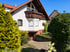 Zweistöckiges weißes Haus mit braunem Holzbalkon und Garage, umgeben von grünen Büschen unter blauem Himmel.