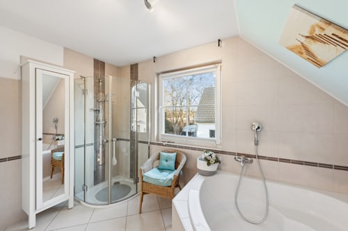 Helles Badezimmer mit weißer Badewanne, Duschkabine, Spiegelschrank, Fenster und Korbsessel.