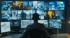 Ein Mann sitzt vor einem großen Bildschirm mit 15 Überwachungsfeeds und drei weiteren Monitoren.
