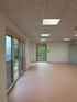 Leerer, heller Raum mit bodentiefen Fenstern, beigem Bodenbelag und Akustikdecke mit quadratischen LED-Leuchten.