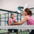 Eine Frau in einem pinkfarbenen Oberteil spielt Padel-Tennis, ein Mann in einem pinkfarbenen T-Shirt steht im Hintergrund.