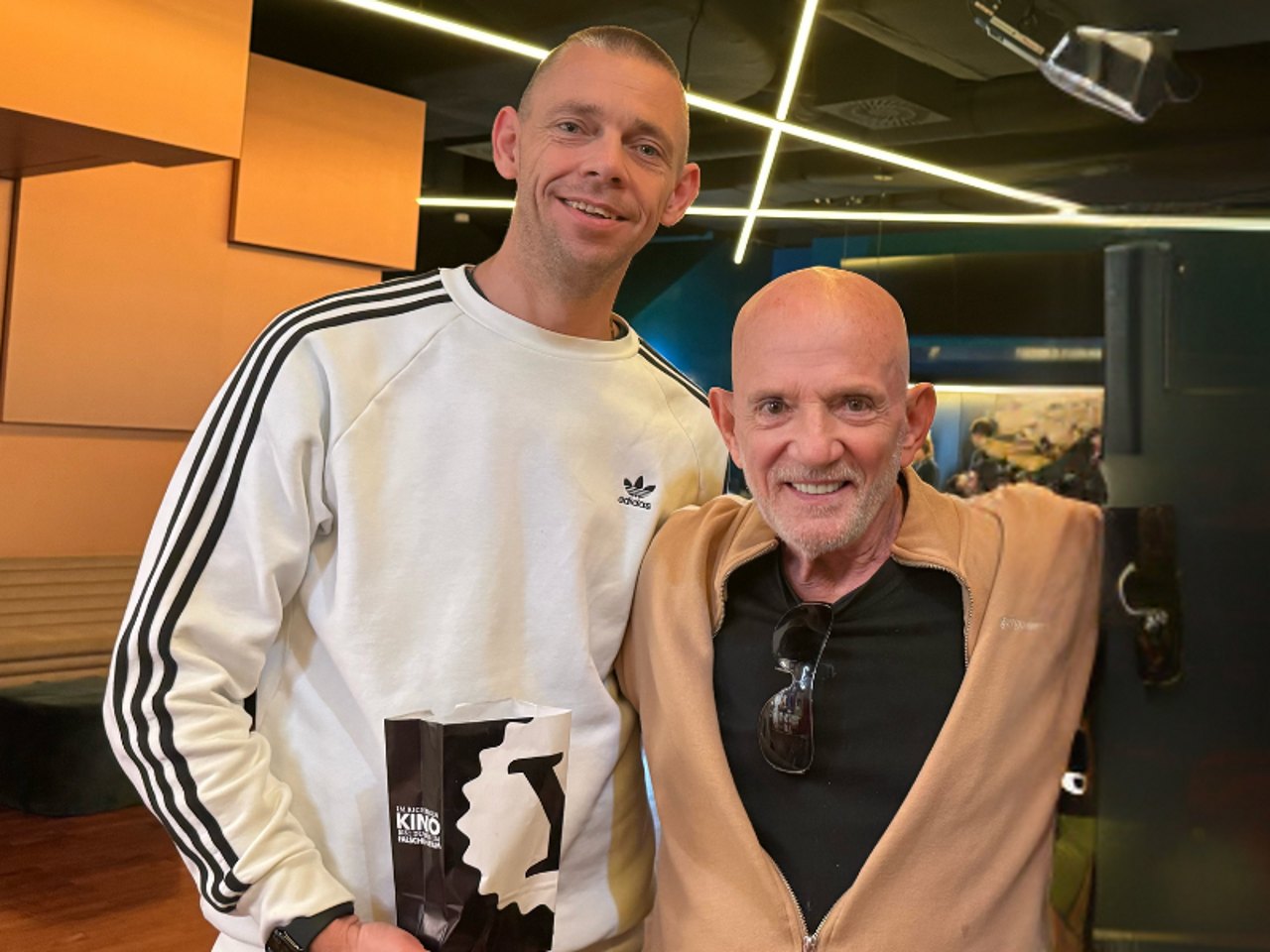 Two men smiling at the camera, one holding a popcorn bag labelled "KINO".